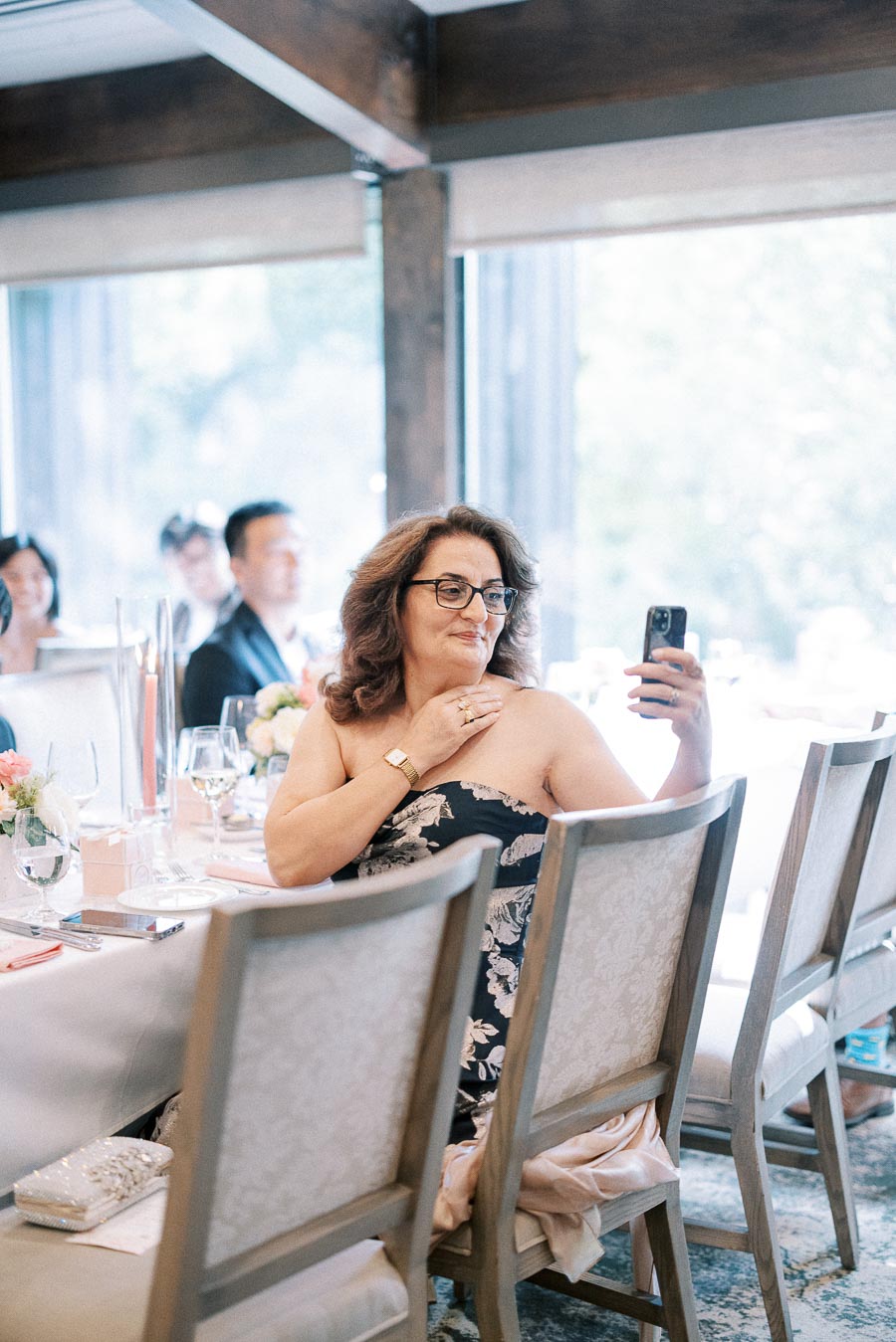 Woman seated at a reception table records with her phone beside candles, flowers, glassware, and bright windows.