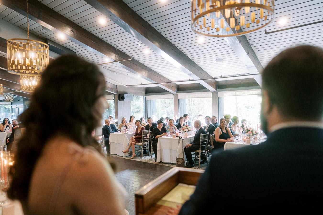 Miffy and Rawle stand blurred in the foreground facing seated reception guests in a bright dining room.