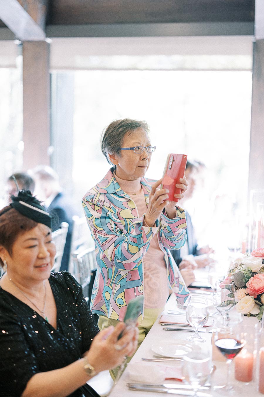 Older wedding guest in a colorful jacket records on a red phone beside a reception table with flowers.