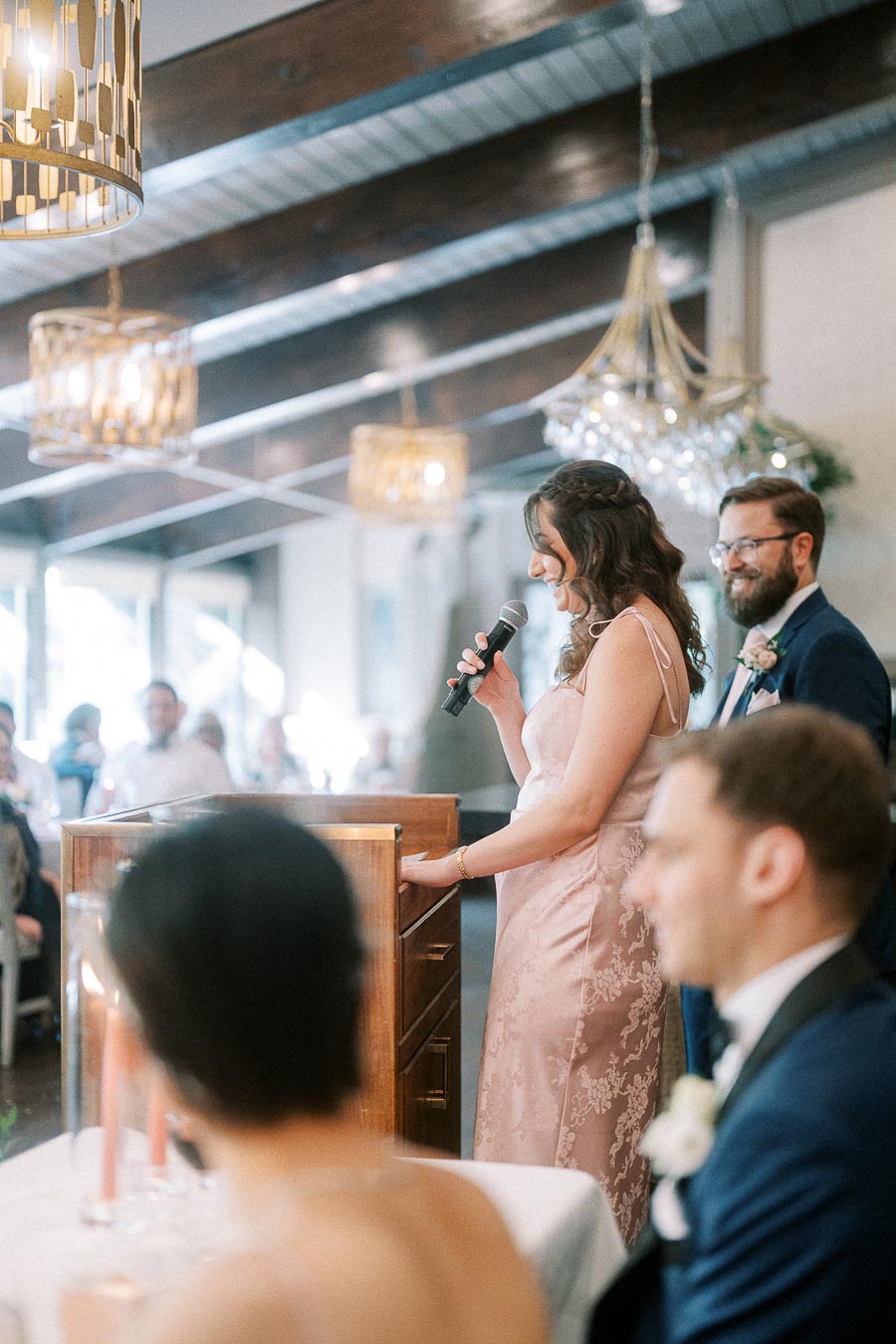 Woman in a pink dress gives a wedding reception speech at a podium while guests listen under chandeliers.