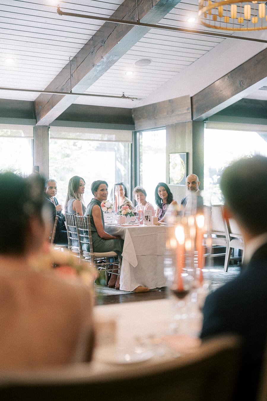 Wedding guests seated at round reception tables in a bright room with wood beams and candles