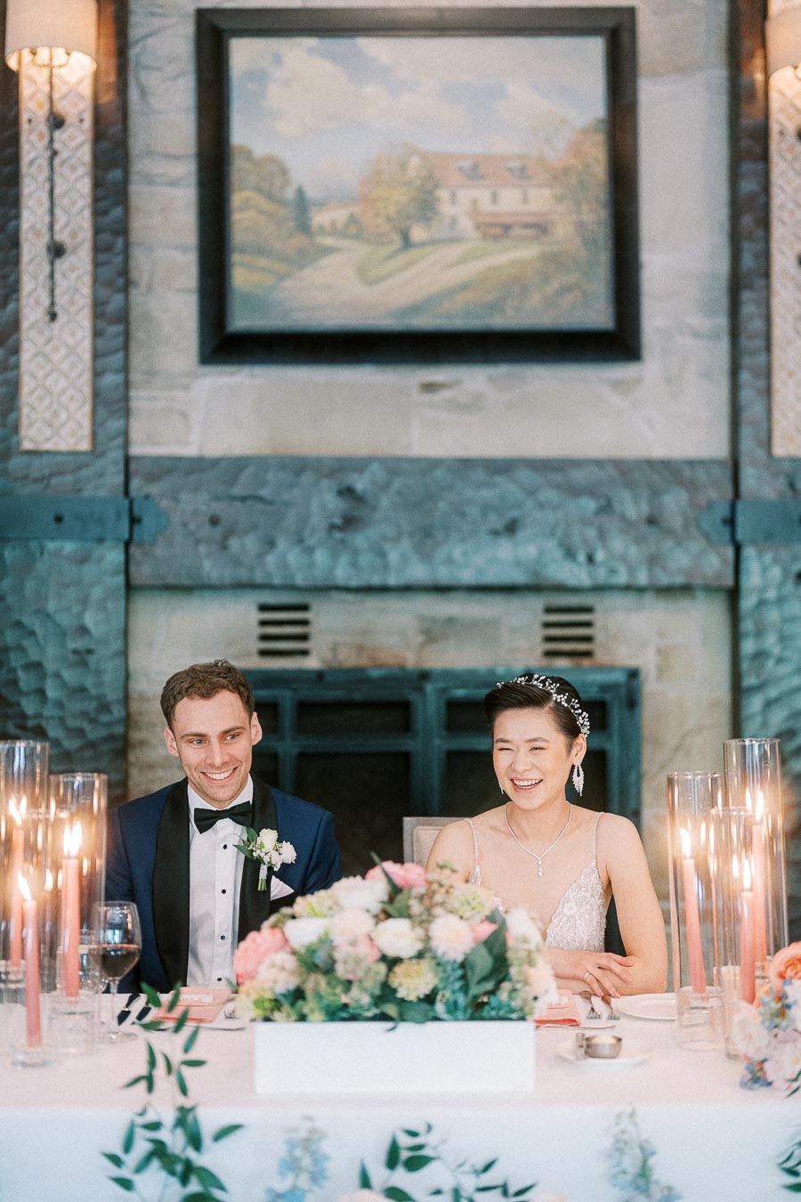 Miffy and Rawle smiling at their candlelit head table beneath a framed painting and stone fireplace
