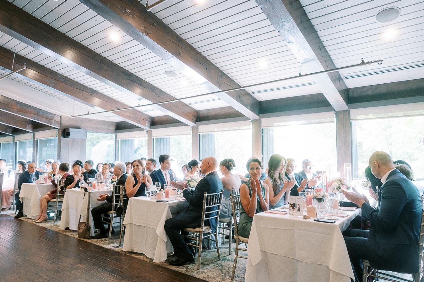 Wedding guests seated at reception tables applaud in a bright room with wood beams and large windows.