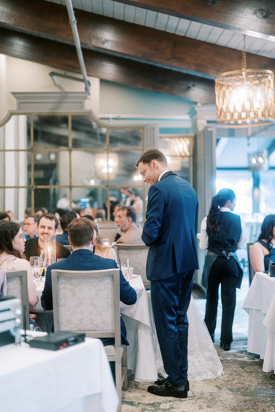 Groom in a navy suit smiles while speaking with seated guests during a bright indoor wedding reception.