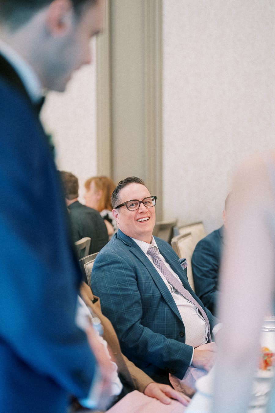 Smiling wedding guest in a blue suit seated at a reception table as another guest stands nearby