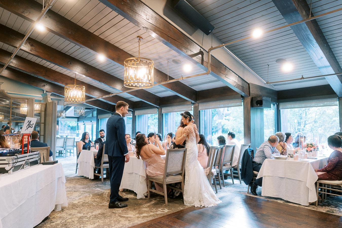 Bride in a lace gown greets a seated guest while the groom stands nearby during an indoor wedding reception.