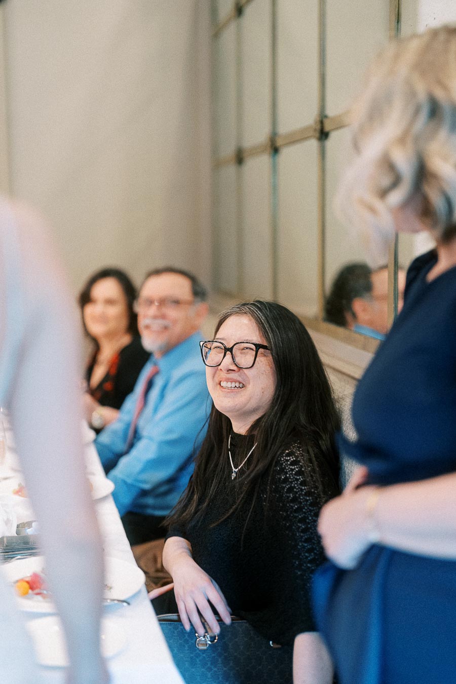 Woman in glasses laughs while seated at a dining table with other wedding guests nearby.