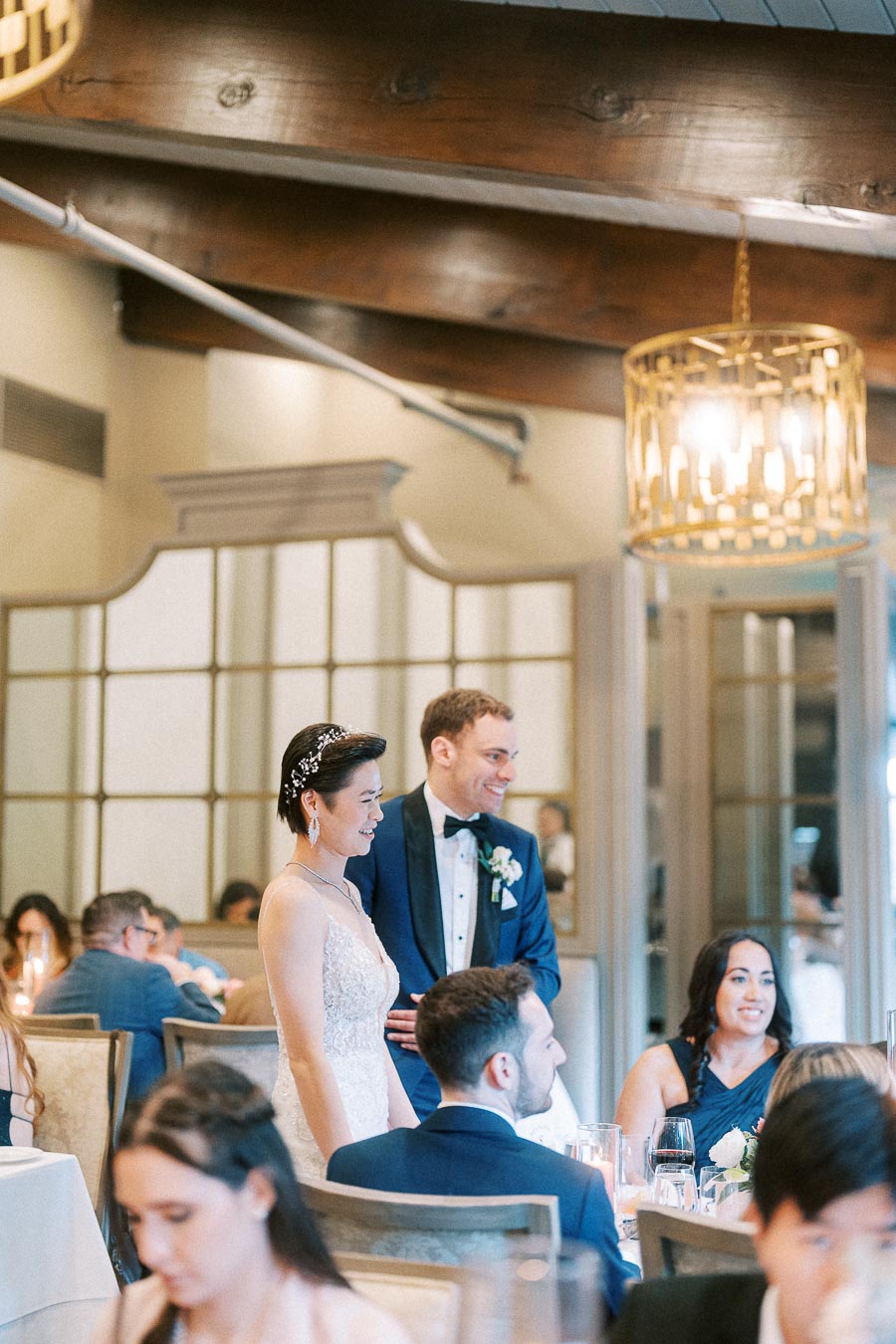 Miffy and Rawle stand smiling among seated wedding guests during their Ancaster Mill reception.