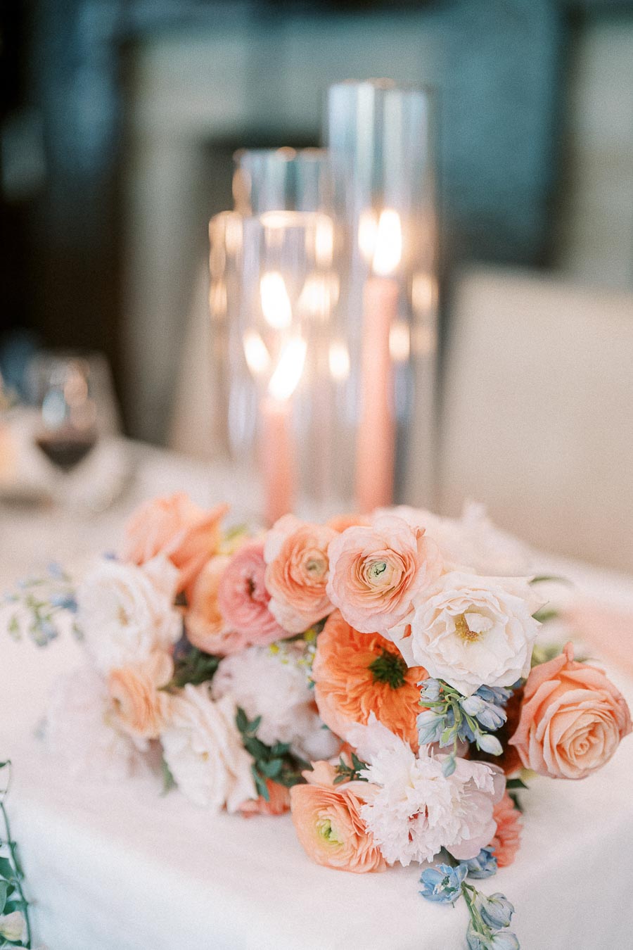Peach and white wedding flowers arranged on a table with glowing taper candles in glass holders behind them.