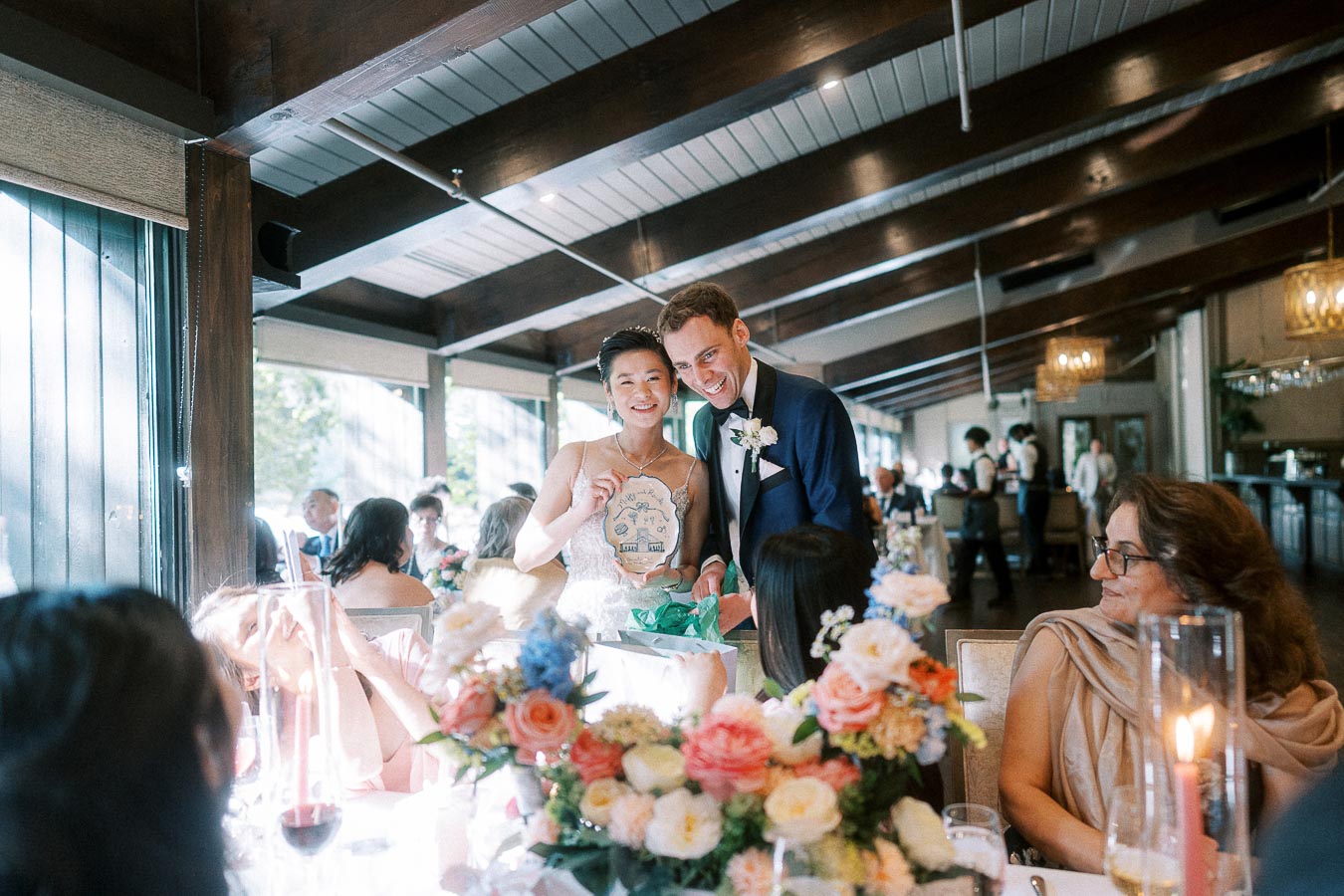 Miffy and Rawle smile beside a reception table while showing a round wedding gift to seated guests.