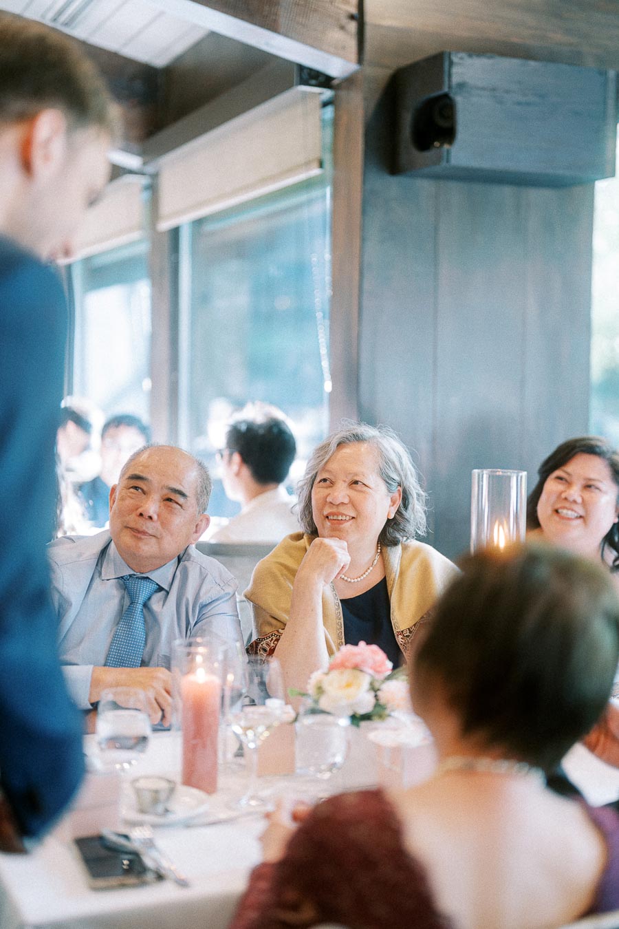 Wedding reception guests at a candlelit table smile up toward a speaker during a speech.