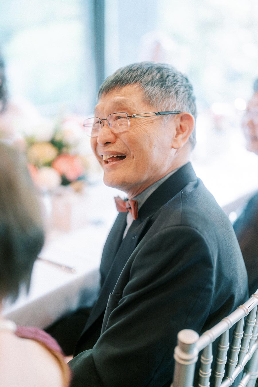 Smiling older man in glasses and a black suit with a red bow tie seated among wedding guests