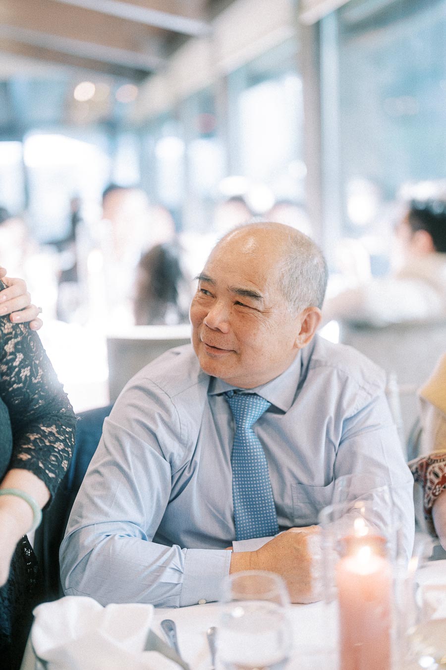Smiling man in a blue tie seated at a candlelit wedding reception table with guests nearby.