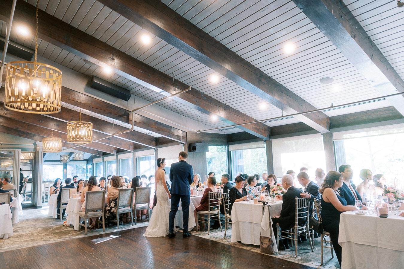 Bride and groom stand among seated wedding guests during a bright reception in a wood-beamed dining room.