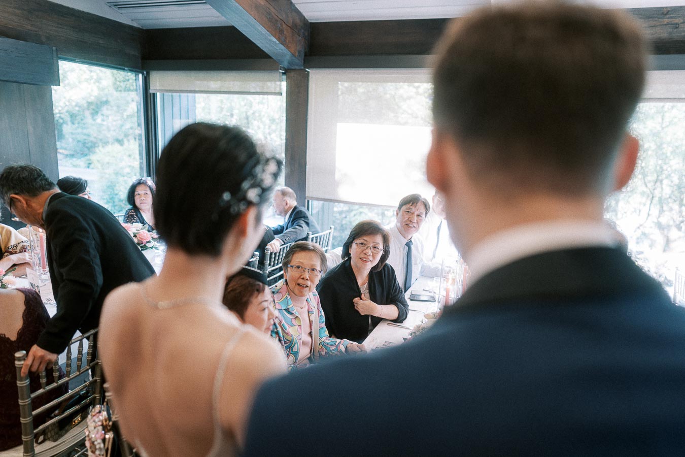 Bride and groom seen from behind greeting seated reception guests in a bright room with large windows.