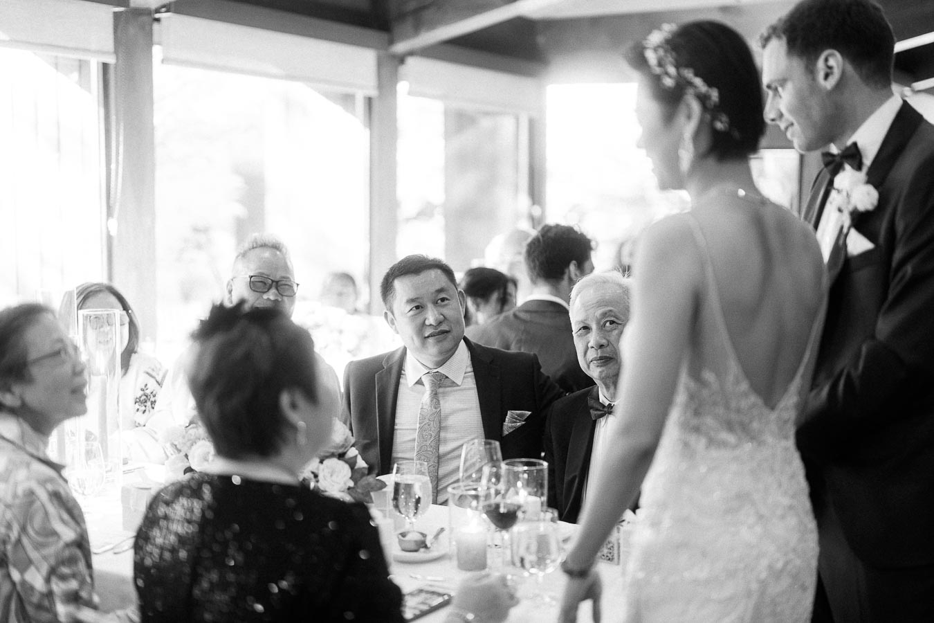 Bride and groom stand beside seated wedding guests during a black-and-white reception conversation.