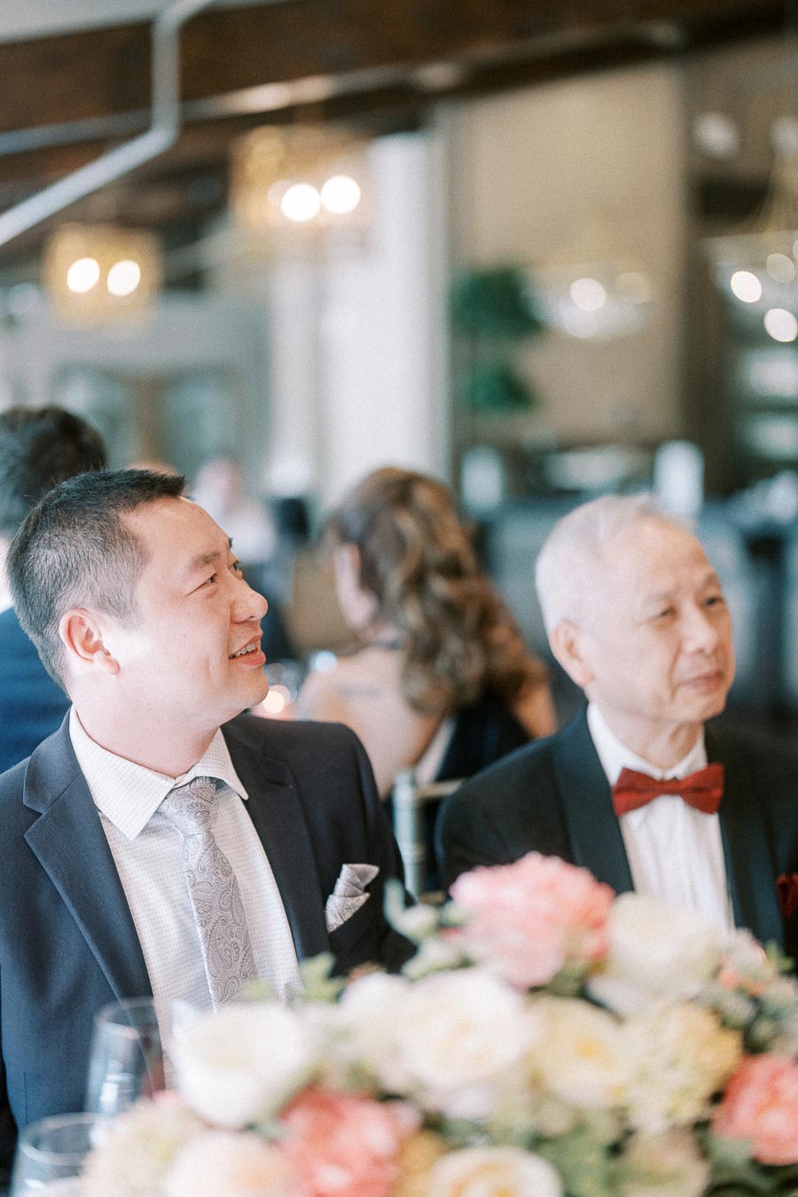 Two suited wedding guests seated at a reception table with pastel floral centrepieces in the foreground.