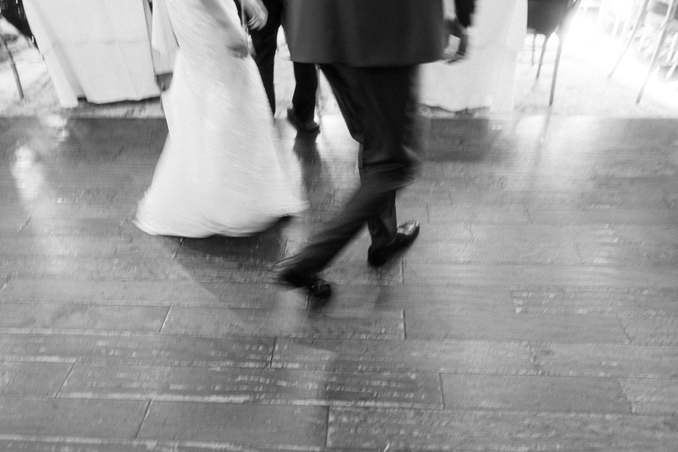 Black and white view of a bride and suited guests walking across a wooden reception floor.