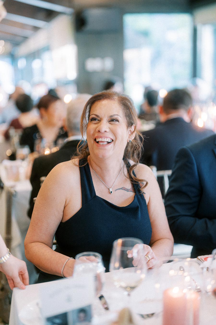 Smiling woman in a black dress seated at a candlelit wedding reception table with guests behind her.