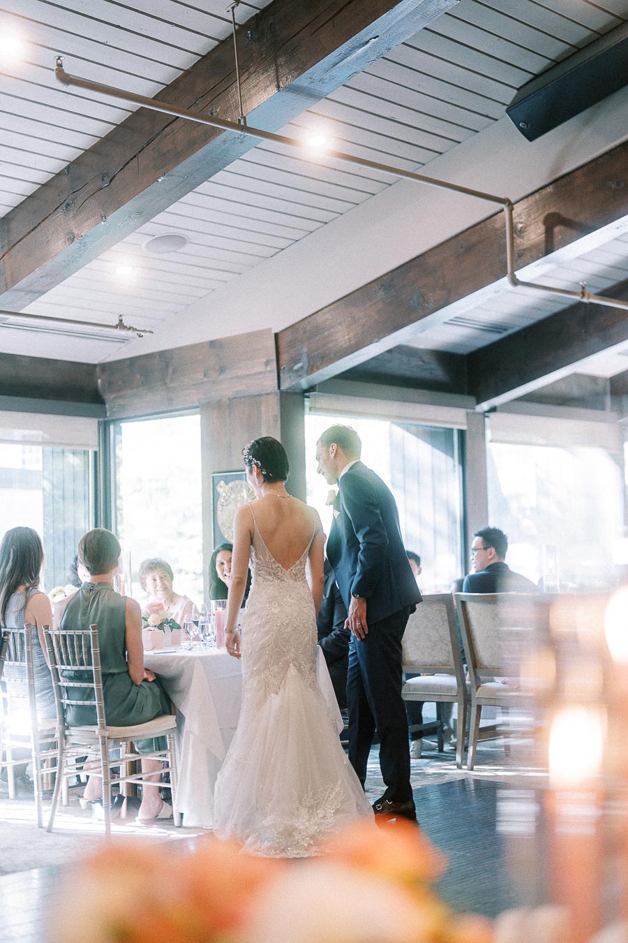 Bride and groom stand beside seated guests during a bright wedding reception with wood beams and tall windows.
