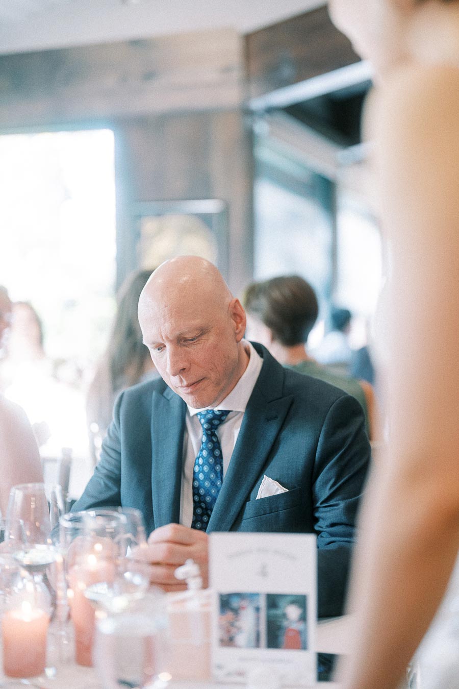 Bald man in a dark suit seated at a candlelit wedding reception table with glassware and a menu nearby.