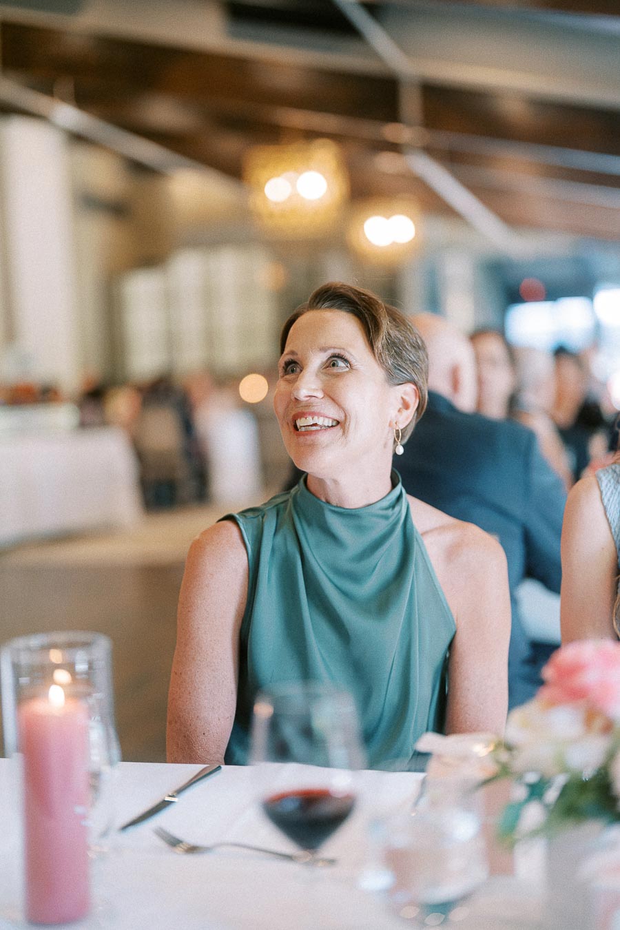 Smiling wedding guest in a teal dress seated at a reception table with candles, flowers, and wine.