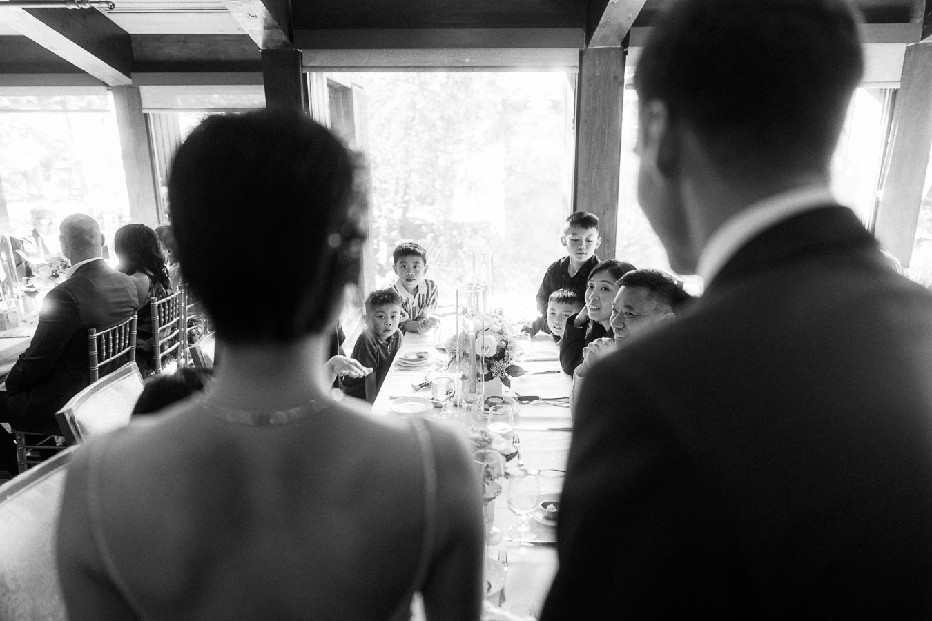 Bride and groom stand beside a reception table as children and guests look toward them.