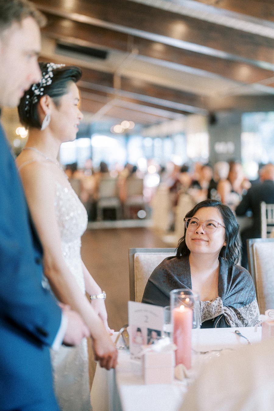 Bride and groom stand beside a reception table while a seated guest smiles up at them.