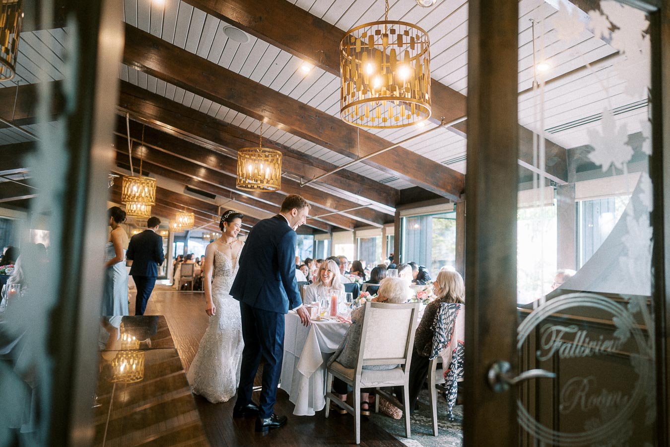 Bride and groom greet seated guests during a bright wedding reception in a wood-beamed dining room.