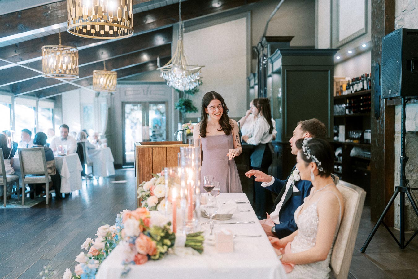 Bridesmaid speaks beside the sweetheart table as the bride and groom listen during the reception.