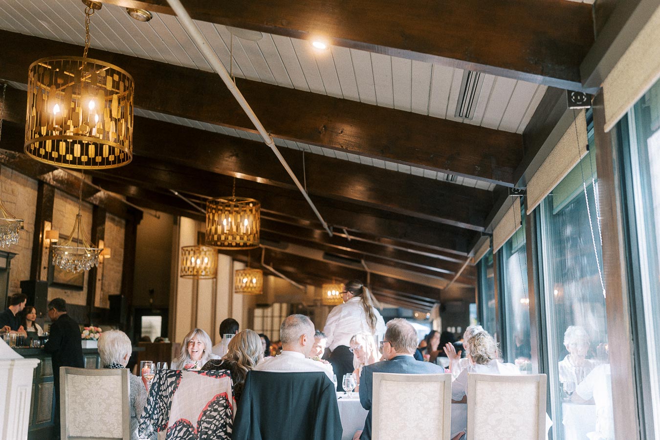 Wedding guests seated at round reception tables beneath chandeliers and wood beams in a bright dining room.
