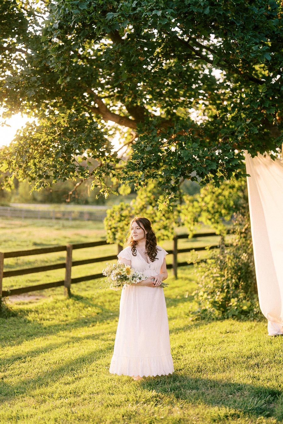Pregnant woman in a white dress holding flowers under a tree at The Green Acres