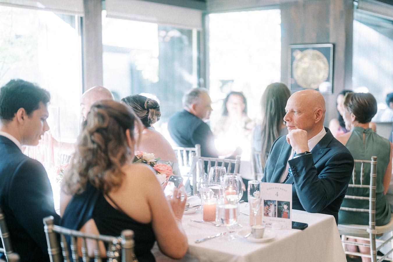 Wedding guests in formal attire sit at a candlelit reception table with wine glasses and a table number card.