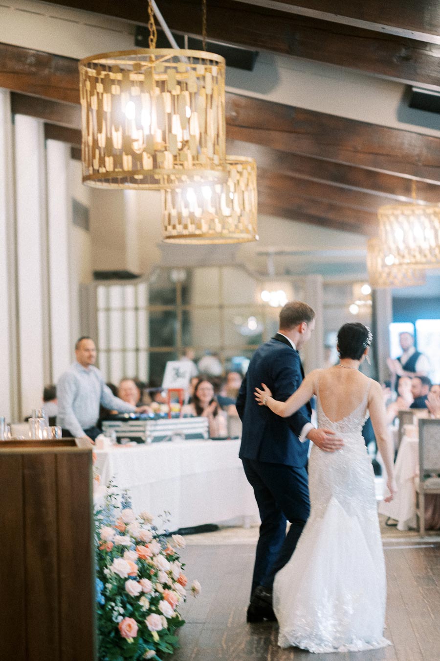 Miffy and Rawle walk arm in arm into their wedding reception beneath gold chandeliers at Ancaster Mill.