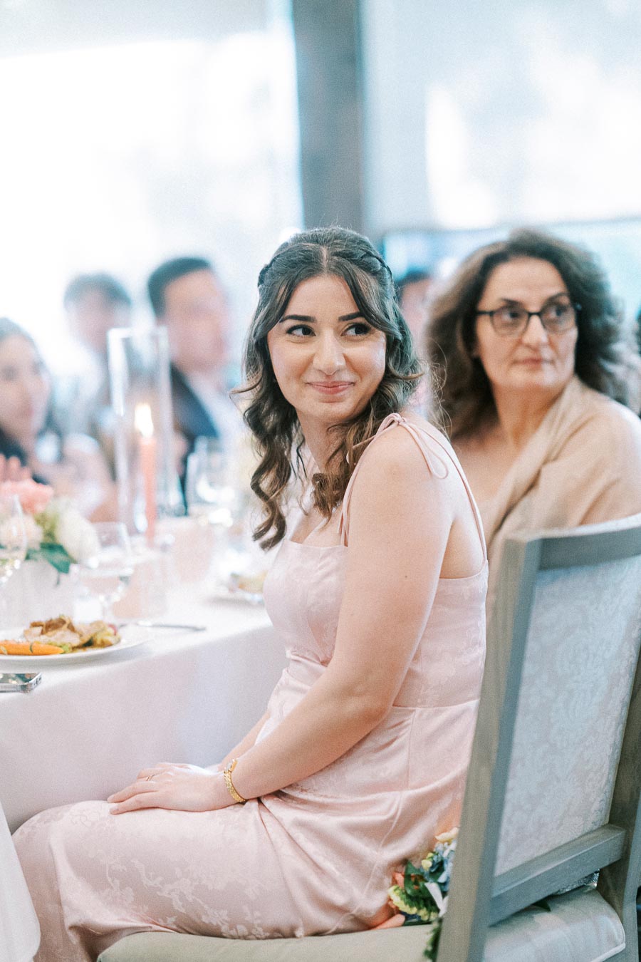 Woman in a pale pink dress seated at a wedding reception table with guests and candles behind her.
