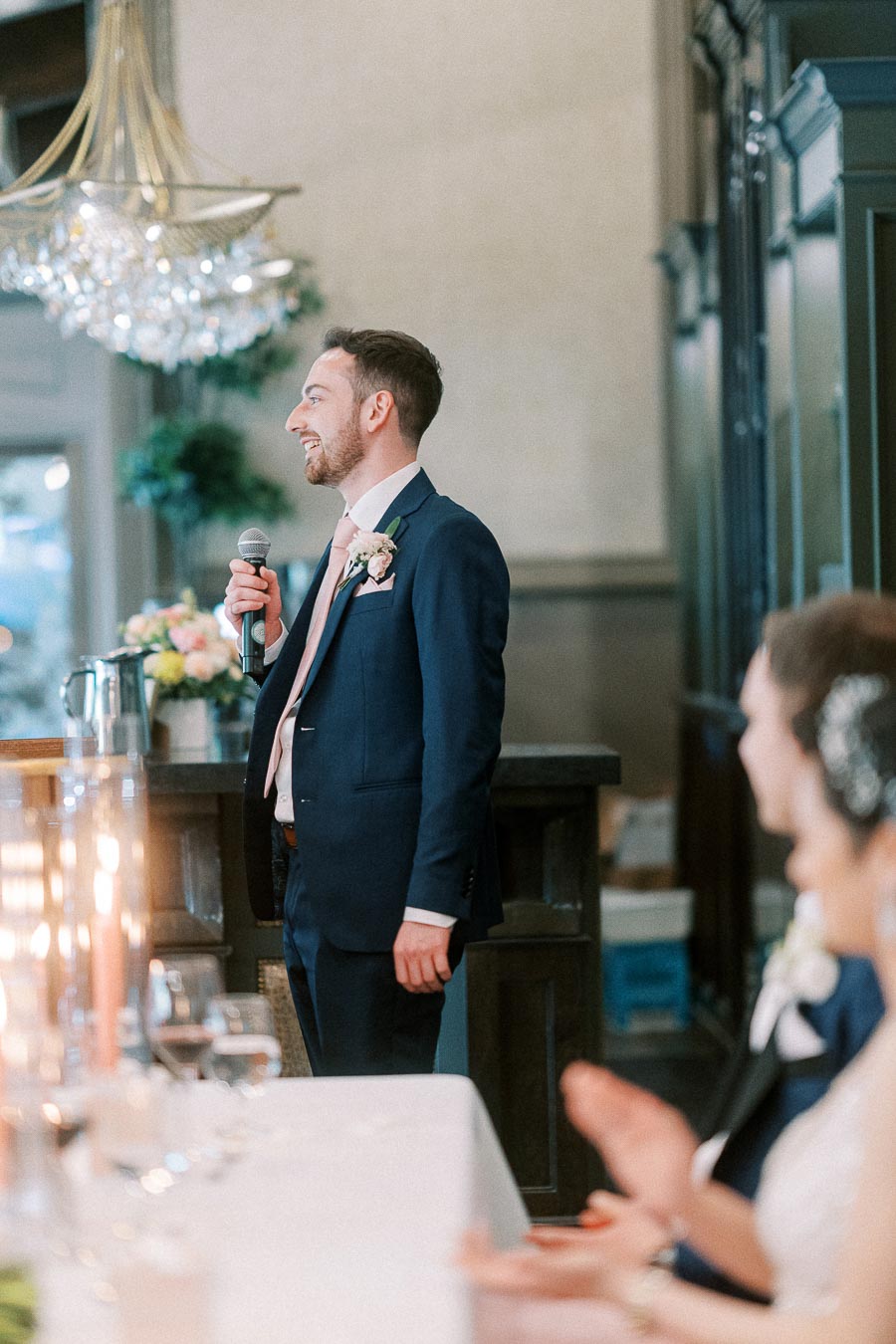 Groomsman in a navy suit smiles while giving a microphone speech beside the wedding head table.