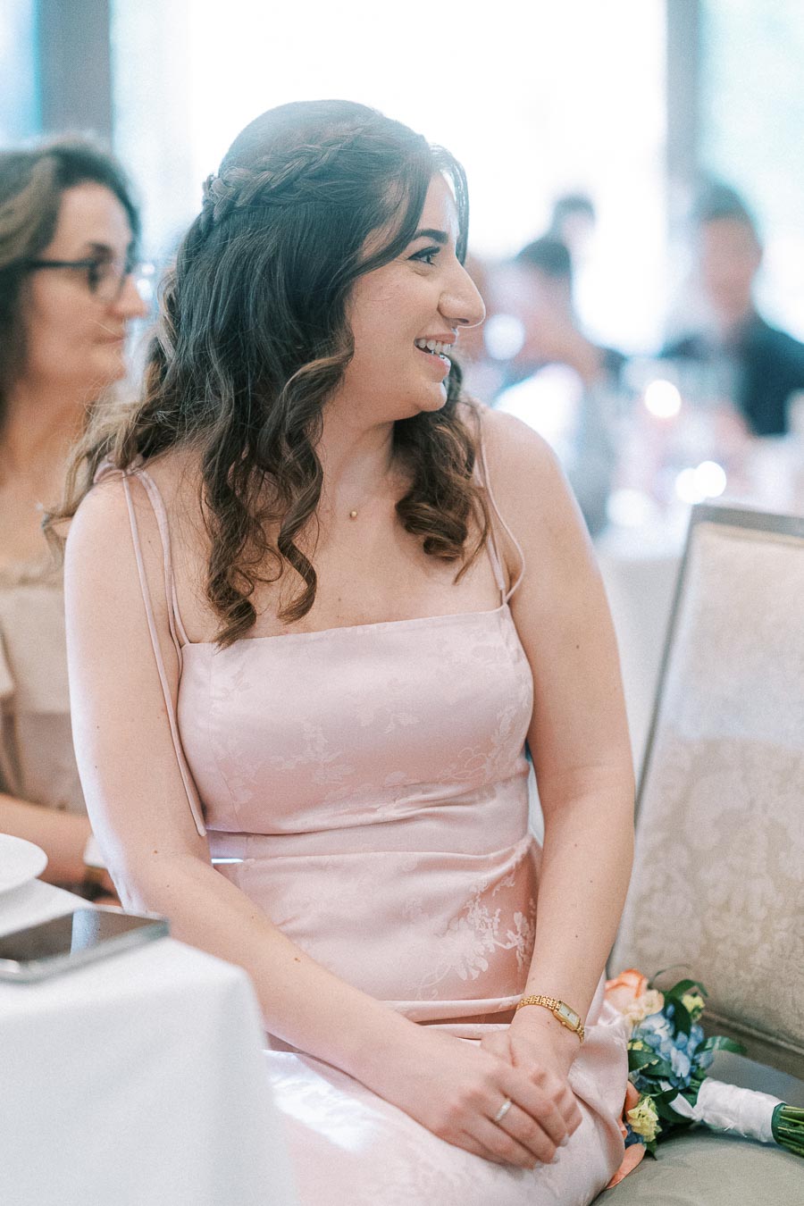 Wedding guest in a pale pink dress smiles while seated at a reception table with a bouquet beside her.