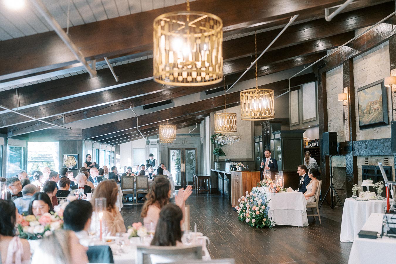 Wedding reception speech in a timber-beamed dining room with guests applauding and the couple seated at the head table.