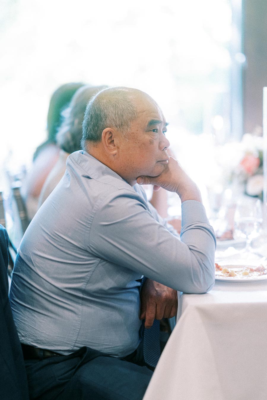 Man in a light blue shirt rests his chin on his hand while seated at a wedding reception table.