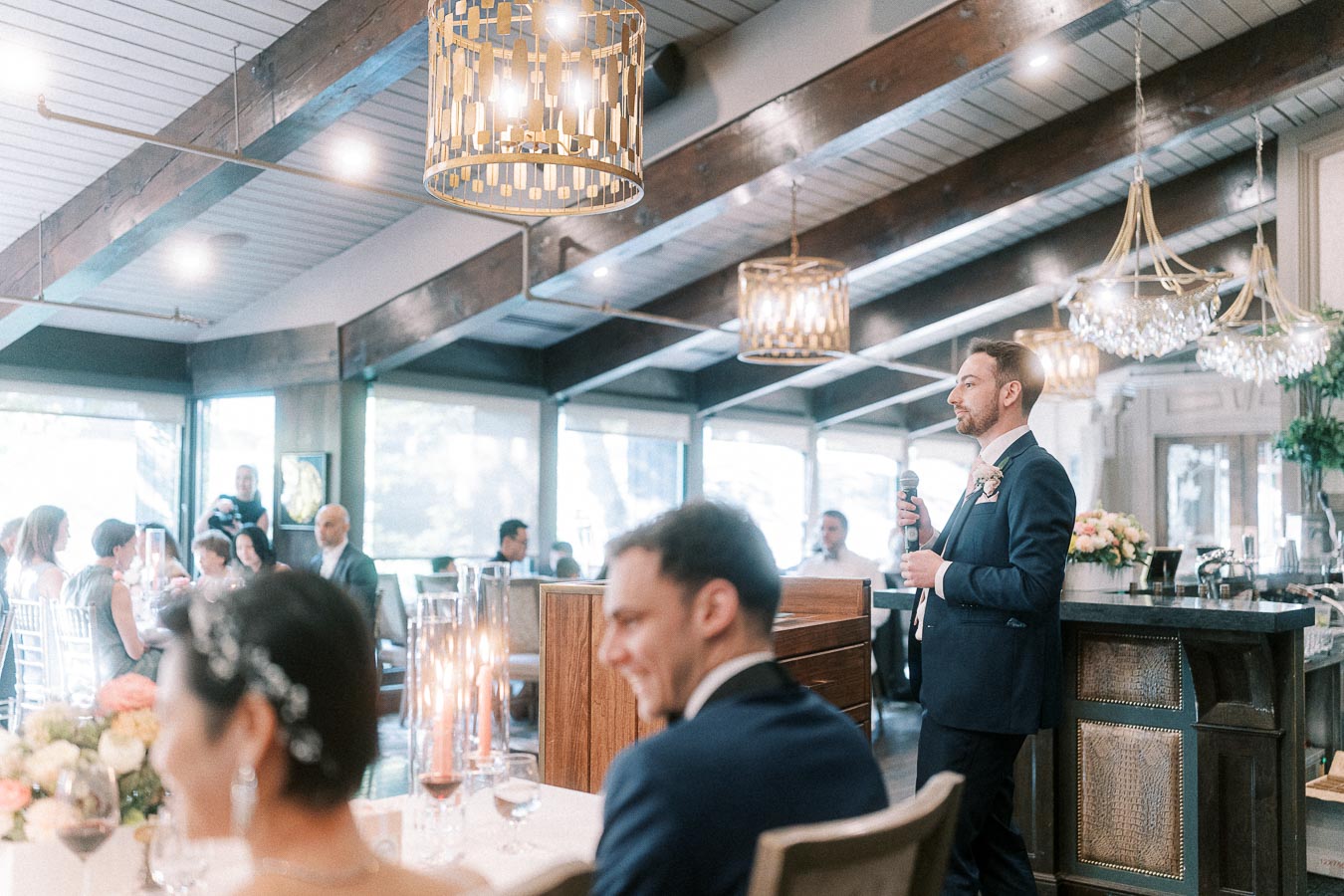 Groomsman gives a reception speech as the couple sits at a head table under chandeliers.