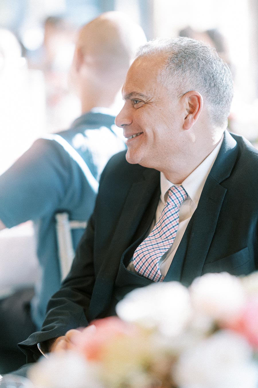 Smiling male wedding guest in a dark suit and pink plaid tie seated at a bright reception table
