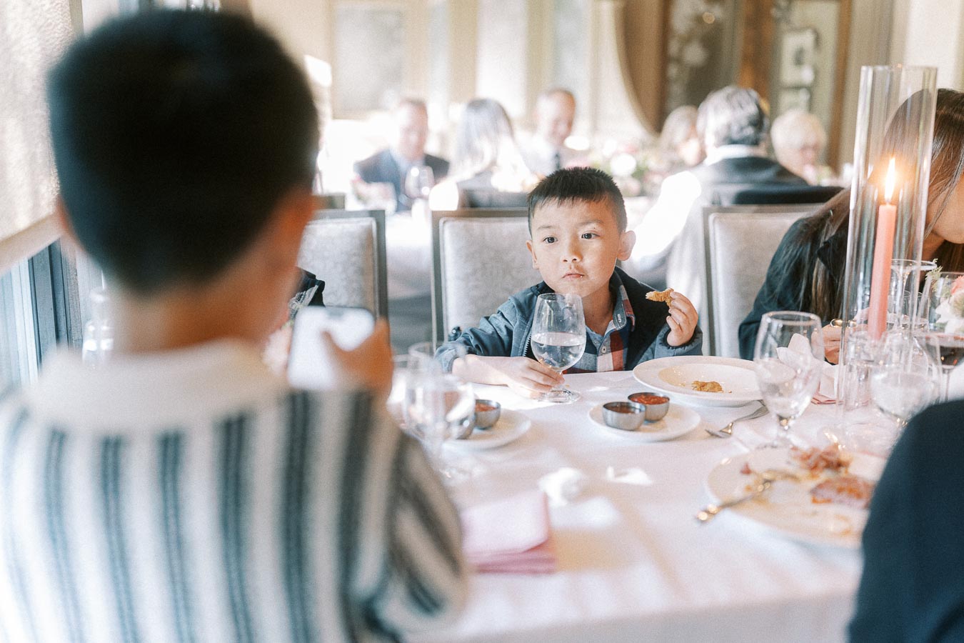 Young boy holding a water glass and food at a candlelit wedding reception table with guests in the background.