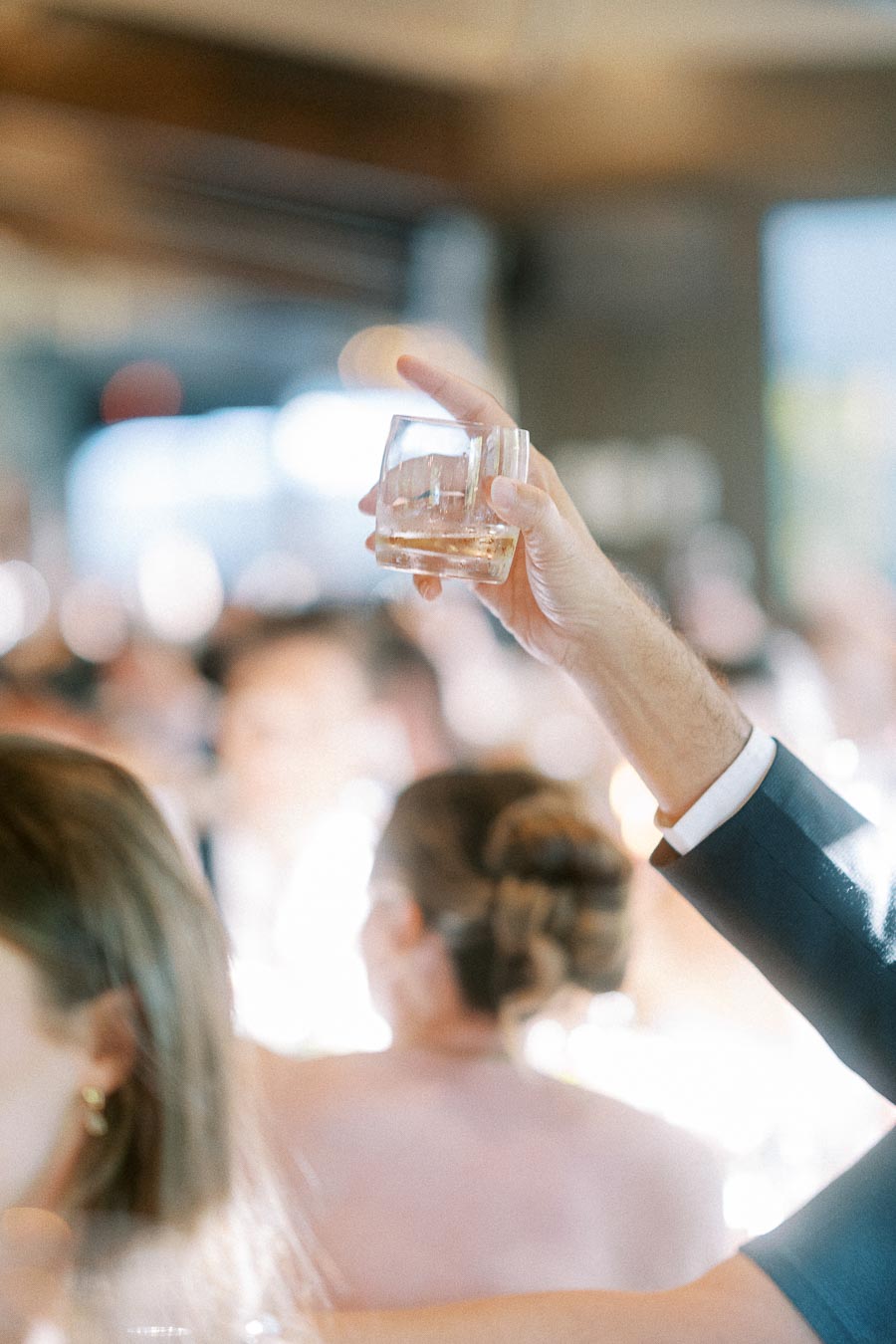 A suited guest raises a short glass of amber drink during a bright wedding reception toast.