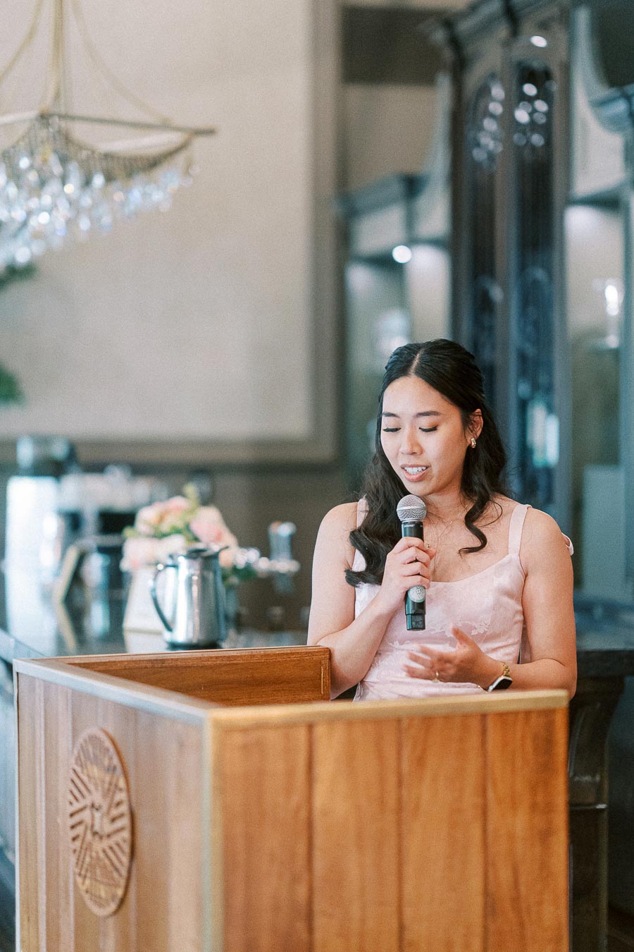 Woman in a blush dress speaks into a microphone behind a wooden podium during a wedding reception.