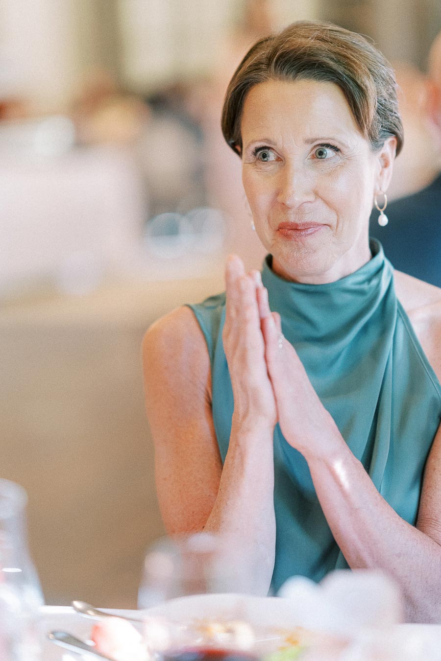 Woman in a teal high-neck dress clasps her hands while seated at a wedding reception table.