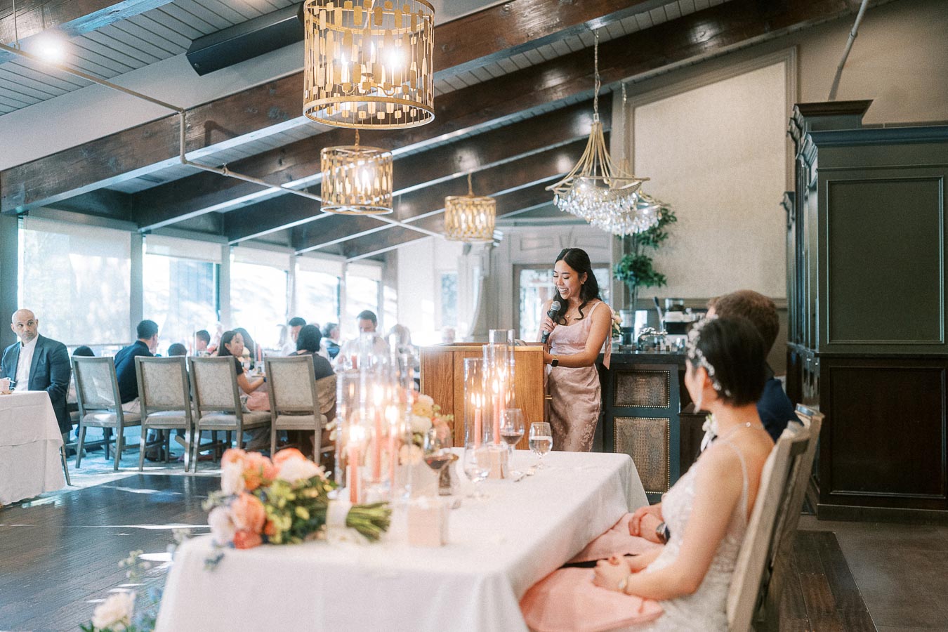 Woman in a pink dress gives a wedding reception speech while the bride listens at the head table.
