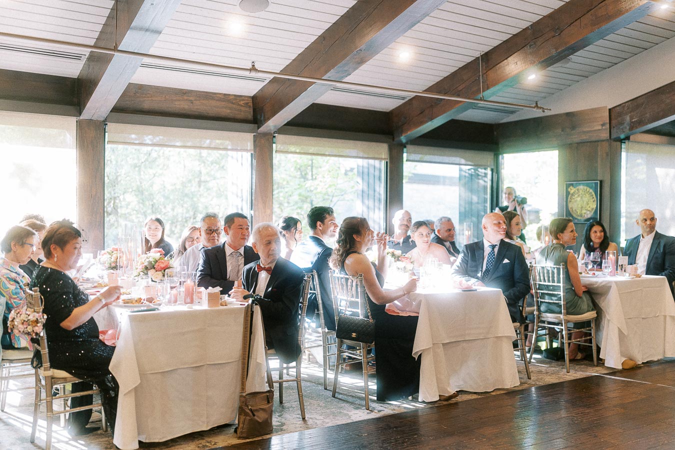 Wedding guests seated at round reception tables in a bright wood-beamed dining room at Ancaster Mill.