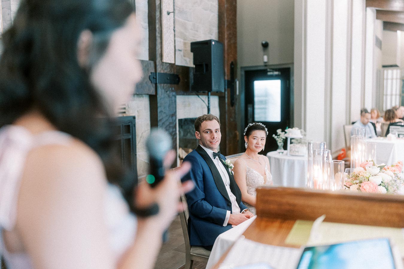 Bride and groom seated at their reception table while a guest gives a speech nearby