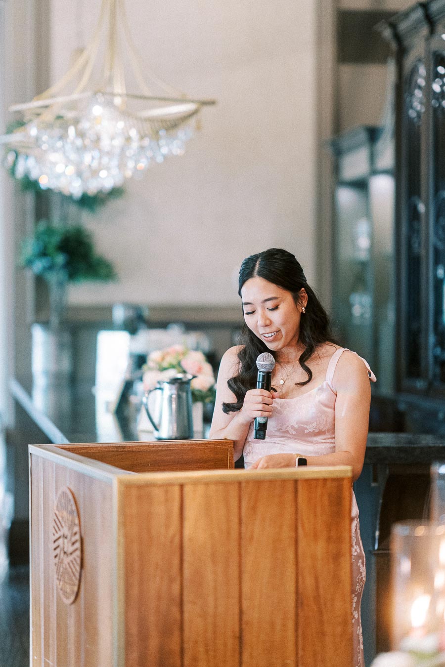 Woman in a pink dress speaks into a microphone behind a wooden podium at a wedding reception.
