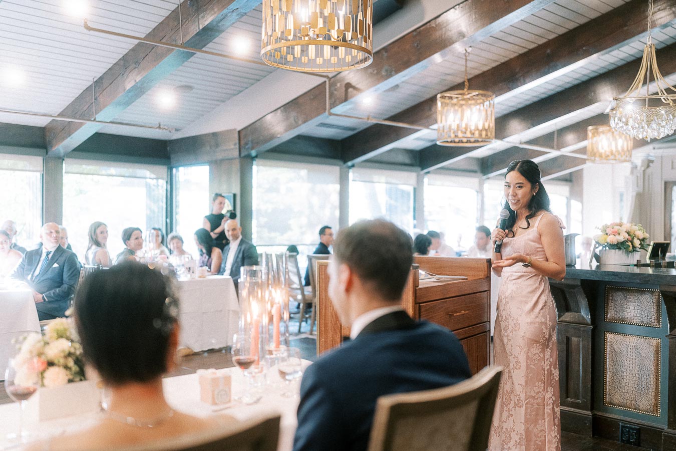 Woman in a pink dress gives a wedding reception speech beside the head table in a bright dining room.